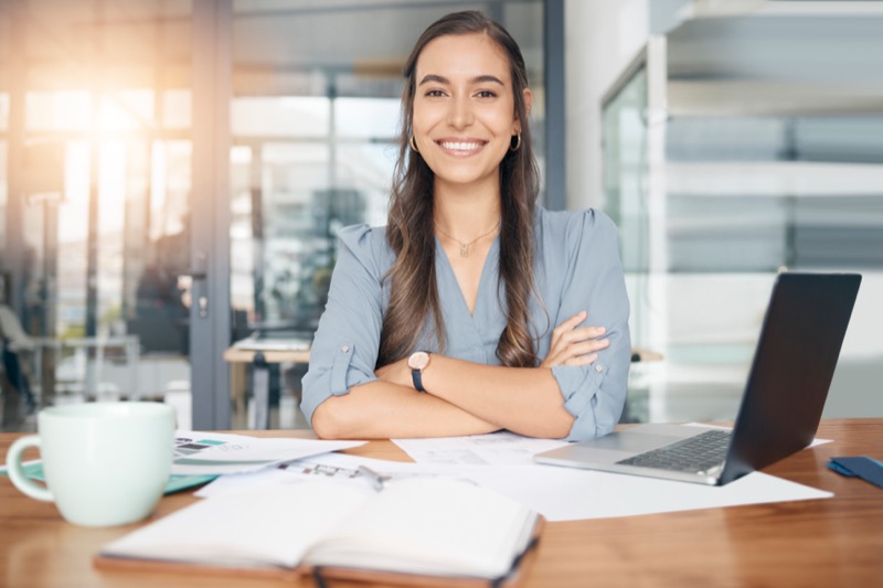 Business woman, portrait and smile at desk in office for paperwork, laptop or administration in Canada. Happy, young or confident female worker with pride planning project at table in startup company.